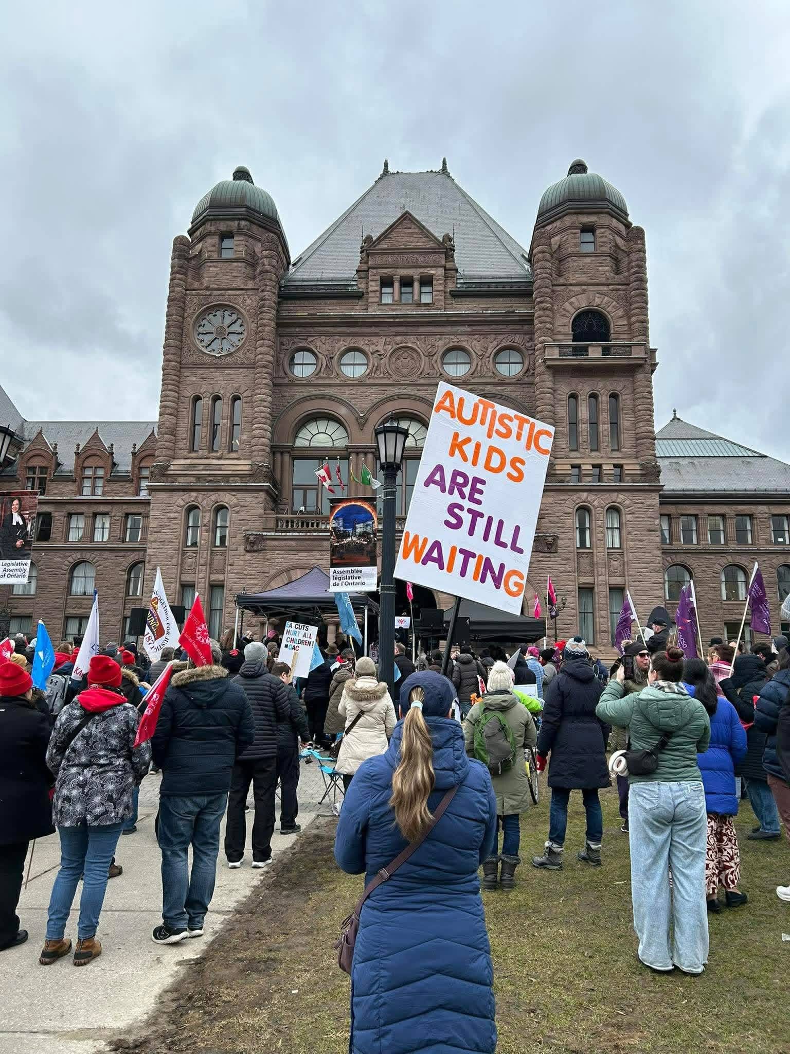 A rally at Queen's Park with a sign reading Autistic kids are still waiting